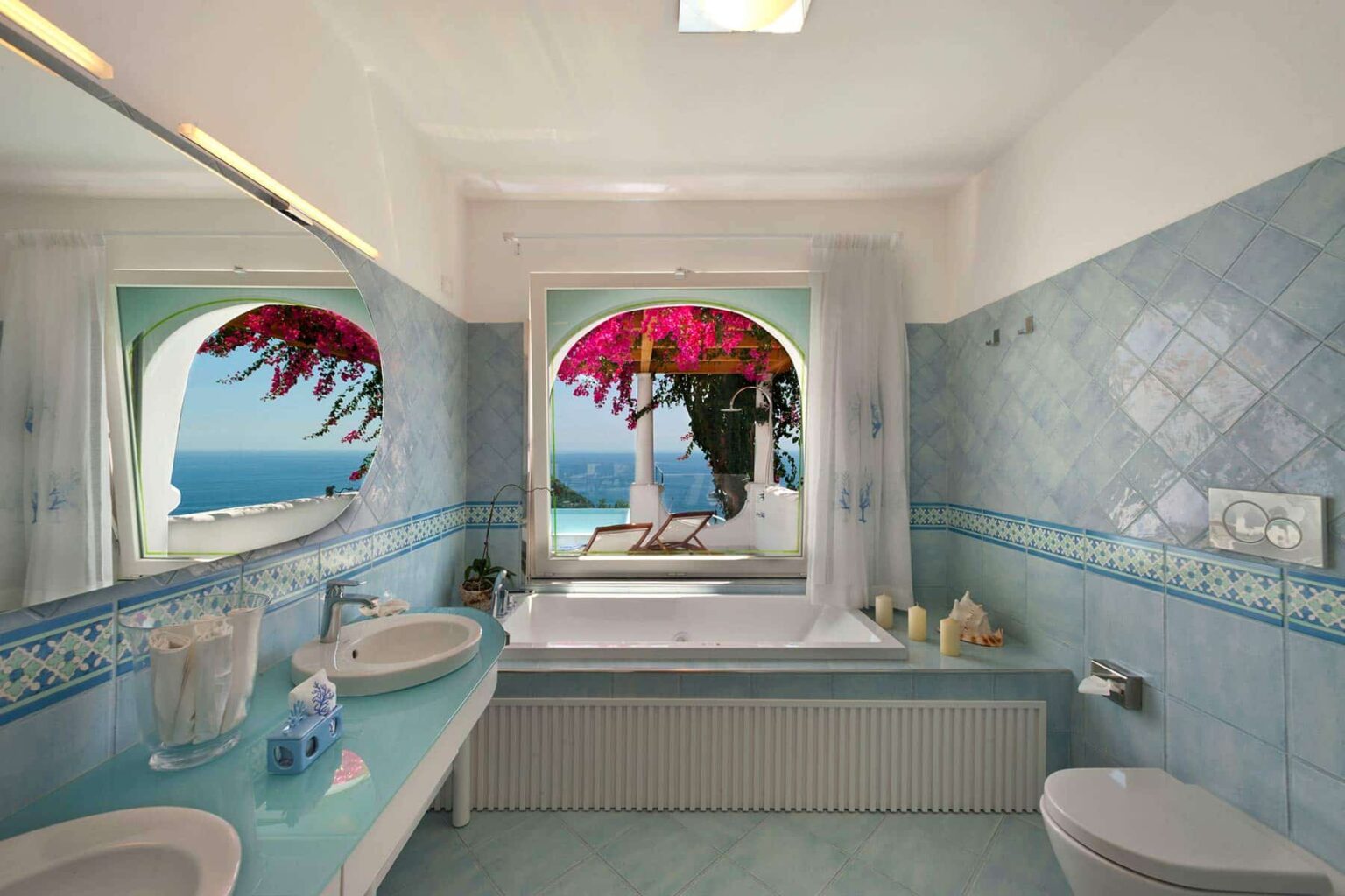 Spacious bathroom with blue tiles, featuring a large bathtub, double sinks, and a picturesque window view of a seaside landscape adorned with pink bougainvillea. | Only on SopranoVillas