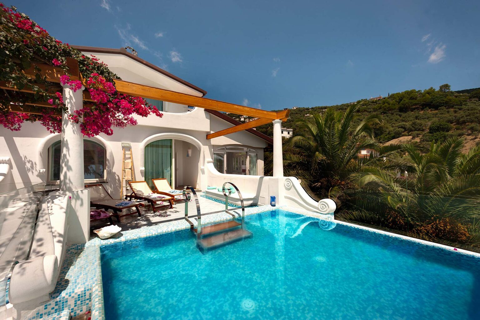 Luxurious villa with a sparkling blue swimming pool surrounded by tropical plants and flowers, featuring a shaded patio area for relaxation and enjoying the scenic hillside views. | Only on SopranoVillas