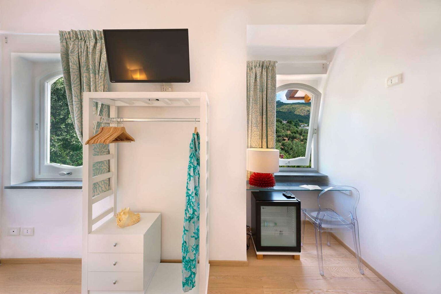 Cozy hotel room featuring a white wardrobe with a hanging dress, a wall-mounted TV, a small desk with a chair, and large windows with green curtains overlooking a scenic view. | Only on SopranoVillas