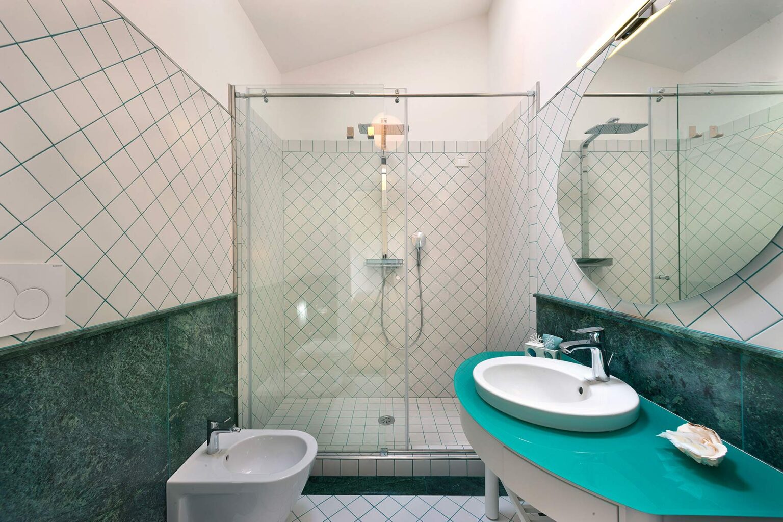 Modern bathroom featuring a glass shower enclosure, a sleek white sink with a turquoise countertop, and a bidet. The walls are tiled with a geometric pattern, and natural light illuminates the space. | Only on SopranoVillas