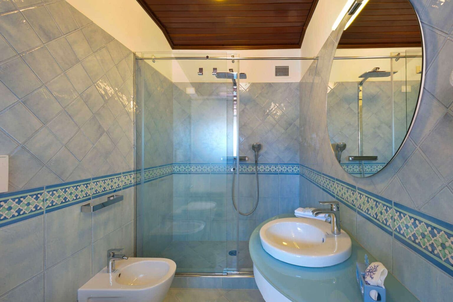 Modern bathroom featuring a glass shower, circular mirror, and stylish blue tile design. The layout includes a bidet and a sleek sink, enhancing the contemporary aesthetic. | Only on SopranoVillas