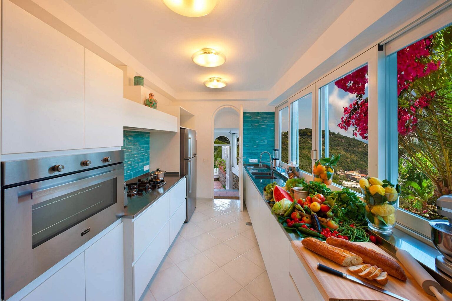 Modern kitchen featuring white cabinetry, stainless steel appliances, and a vibrant turquoise backsplash. A variety of fresh fruits and vegetables are displayed on the countertop, with large windows allowing natural light and a view of blooming flowers outside. | Only on SopranoVillas