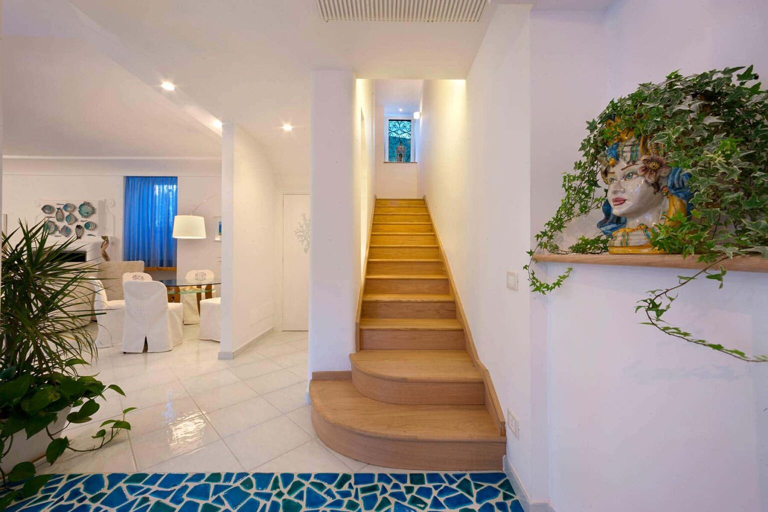 Modern interior design featuring a bright hallway with wooden stairs leading to an upper level. The space is adorned with greenery and a decorative wall piece, complemented by soft lighting and a glimpse of a stylish living area in the background. | Only on SopranoVillas