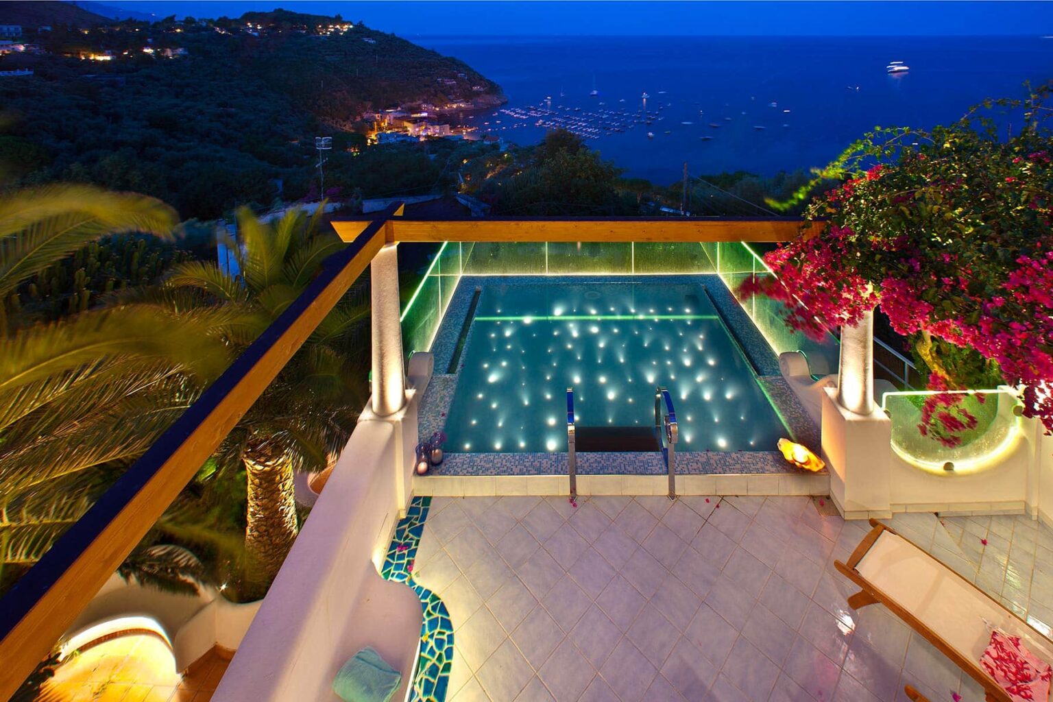 Luxurious villa with a stunning infinity pool overlooking the ocean at dusk, featuring ambient lighting and vibrant tropical flowers. | Only on SopranoVillas
