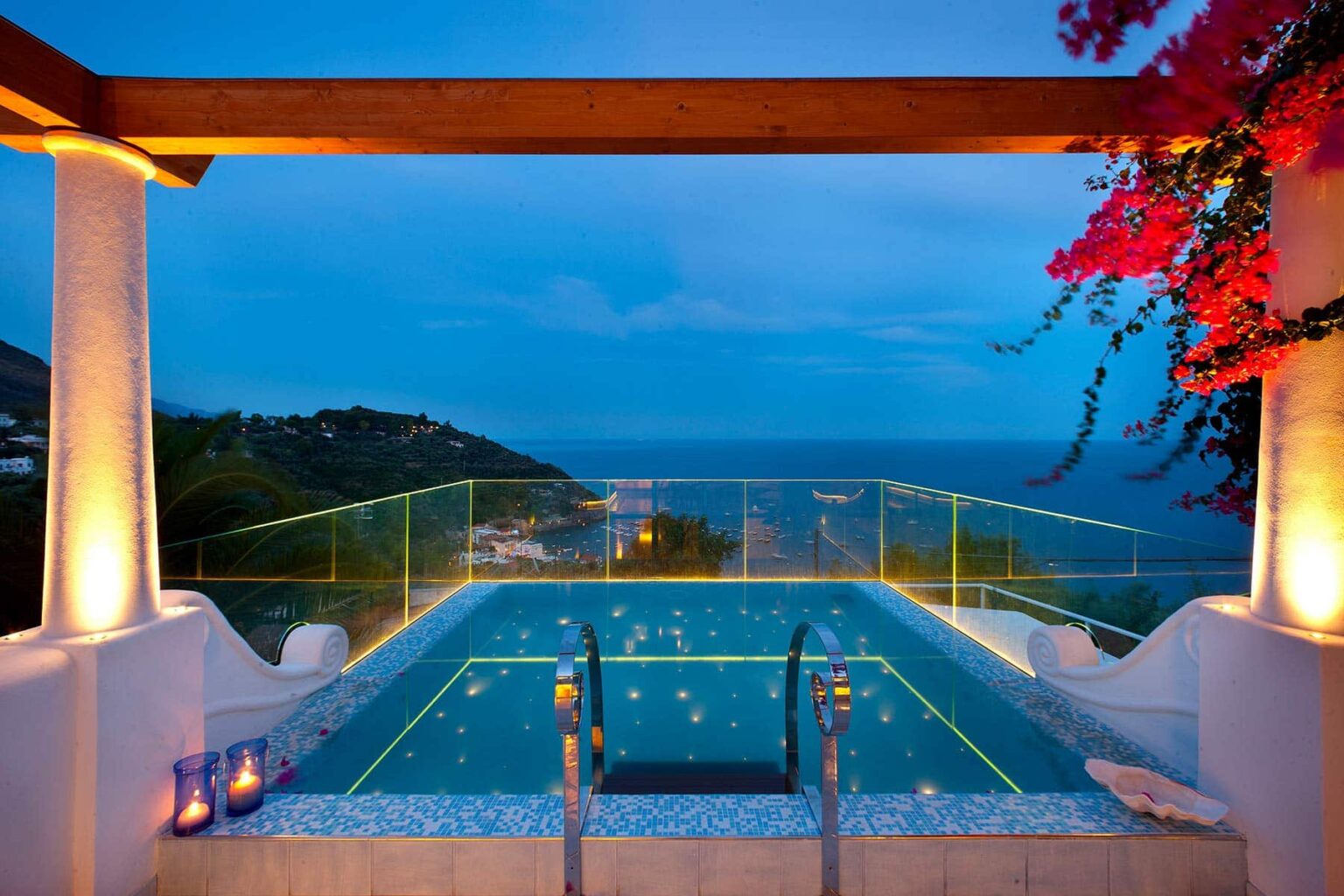 Luxurious infinity pool with ocean view at twilight, surrounded by elegant lighting and vibrant bougainvillea, creating a serene atmosphere for relaxation. | Only on SopranoVillas