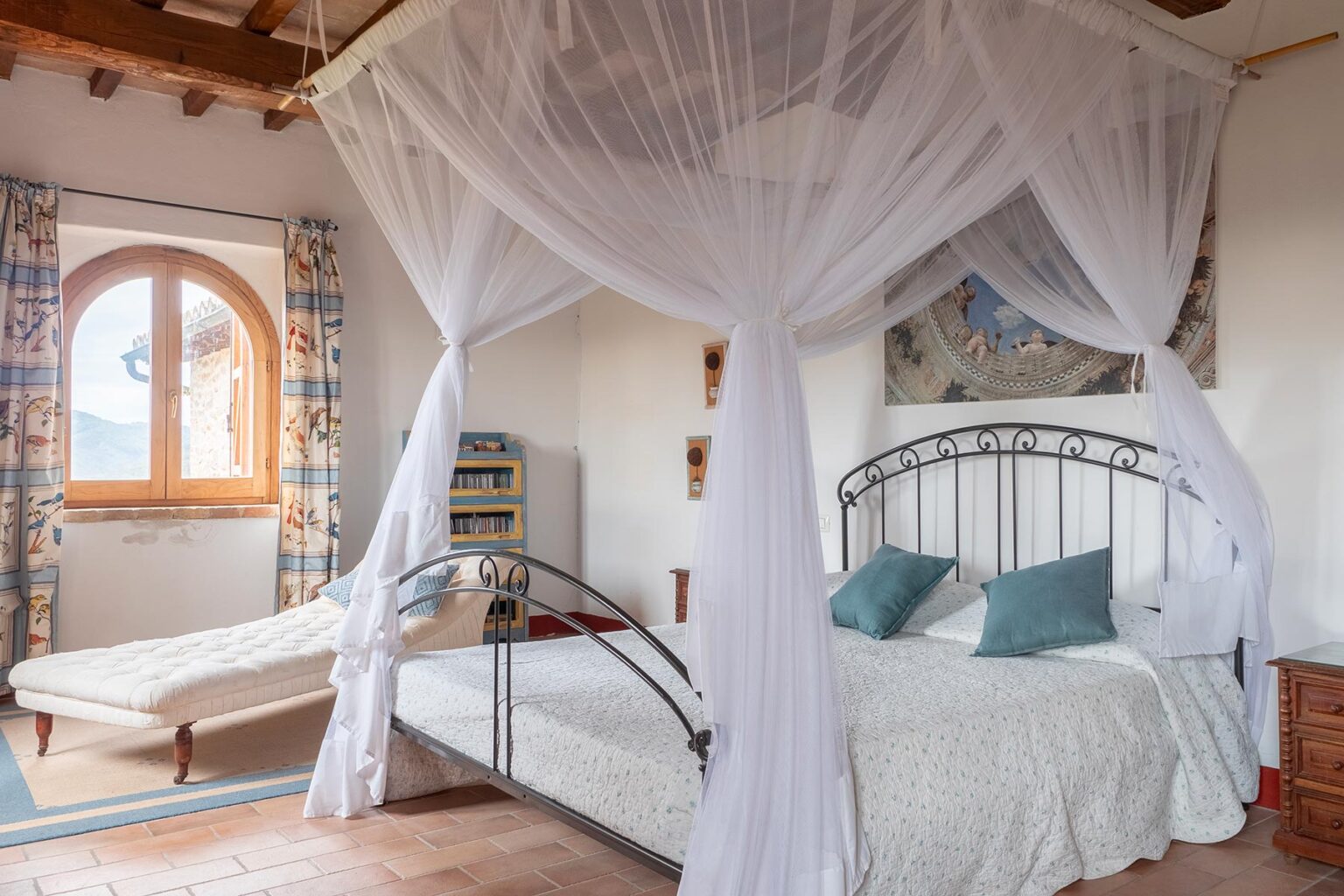 Cozy bedroom featuring a wrought iron bed with a white canopy, soft blue pillows, and a light-colored quilt. A chaise lounge sits beside a large arched window with patterned curtains, creating a bright and inviting atmosphere. | Only on SopranoVillas