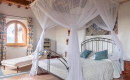Cozy bedroom featuring a wrought iron bed with a white canopy, soft blue pillows, and a light-colored quilt. A chaise lounge sits beside a large arched window with patterned curtains, creating a bright and inviting atmosphere.