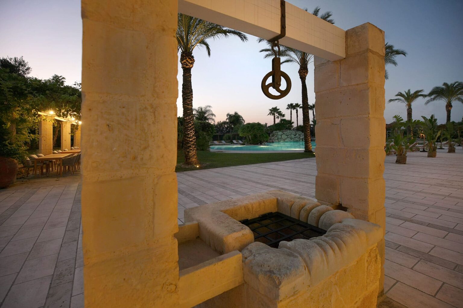 Stone well with a pulley system, surrounded by palm trees and a serene pool area, illuminated by soft evening light. Ideal for outdoor landscaping and architectural design inspiration. | Only on SopranoVillas