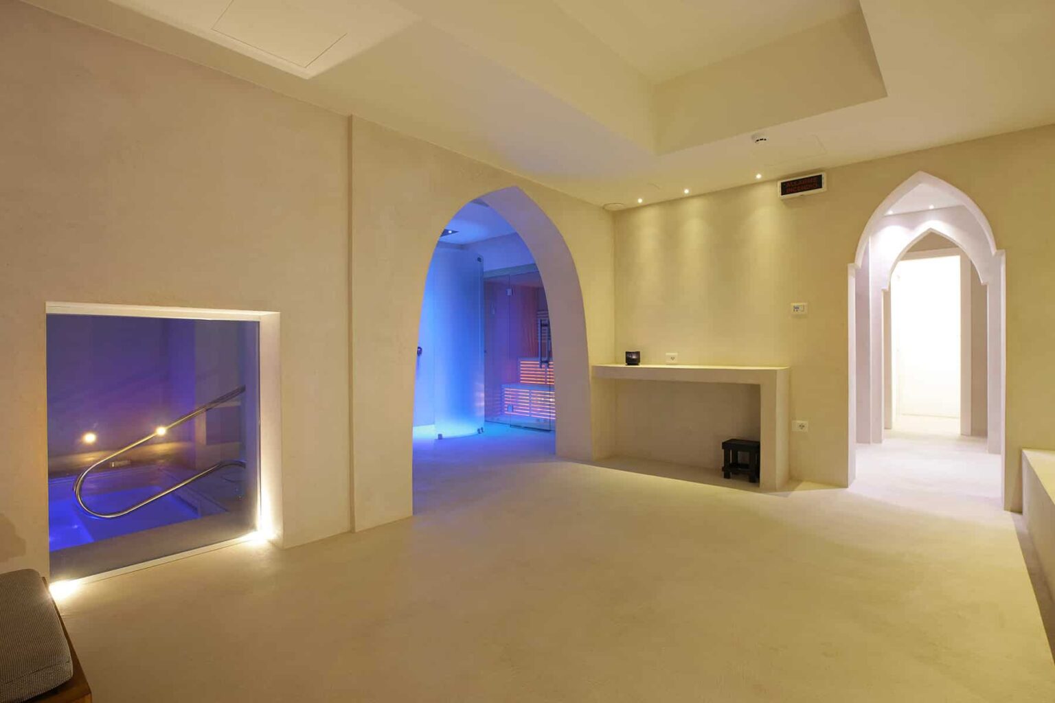 Modern spa interior featuring arched doorways, soft ambient lighting, and a serene atmosphere. The space includes a relaxation area with subtle blue lighting and a wellness treatment room, designed for tranquility and rejuvenation. | Only on SopranoVillas