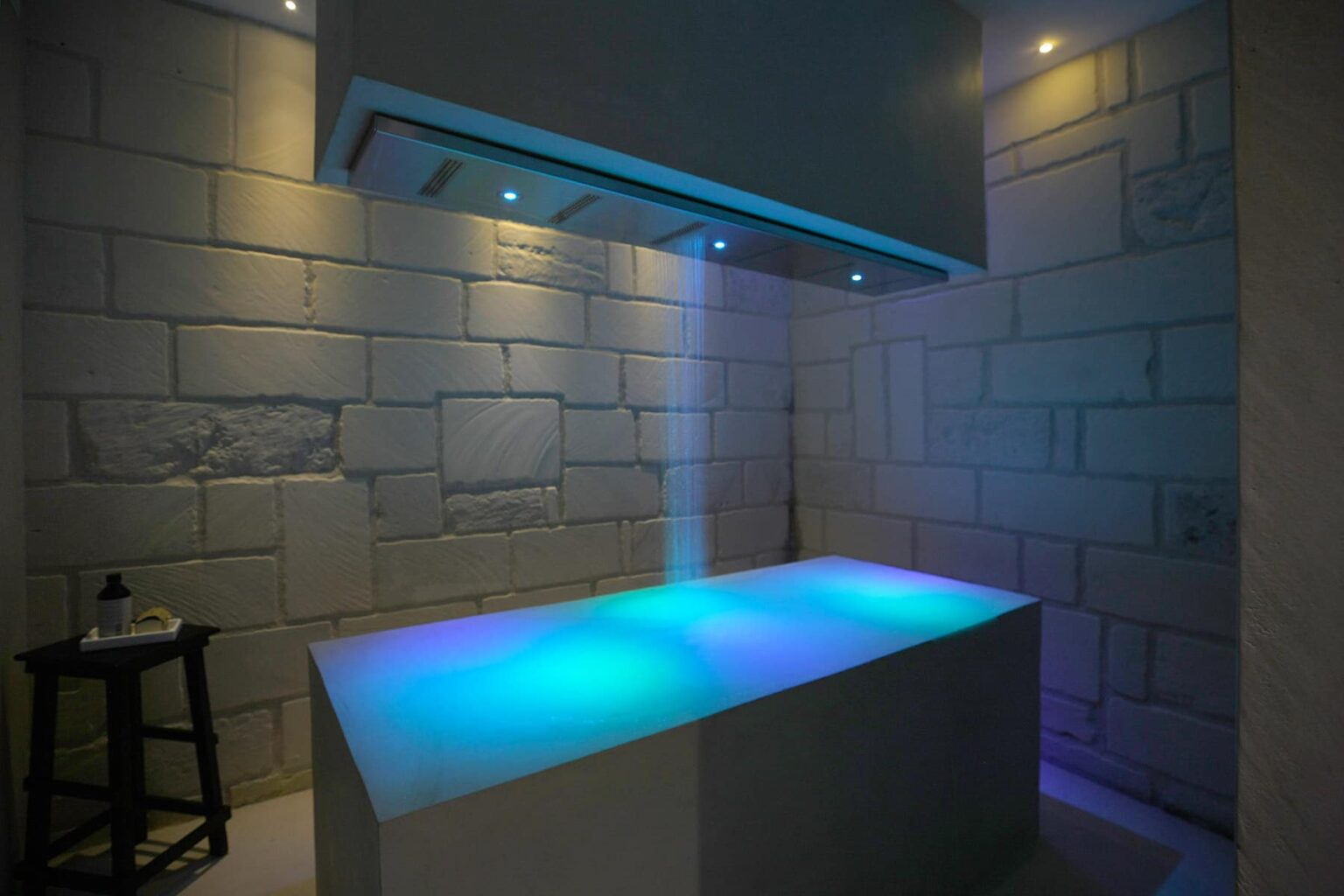 Modern spa treatment room featuring a stone wall design, illuminated with soft blue and purple lighting, and a sleek massage table. Perfect for relaxation and wellness experiences. | Only on SopranoVillas