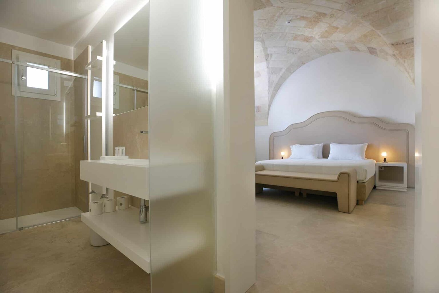 Modern hotel room featuring a spacious bedroom with a large bed, ambient lighting, and an arched ceiling. The adjoining bathroom includes a sleek shower and minimalist design elements, creating a stylish and comfortable atmosphere for guests. | Only on SopranoVillas