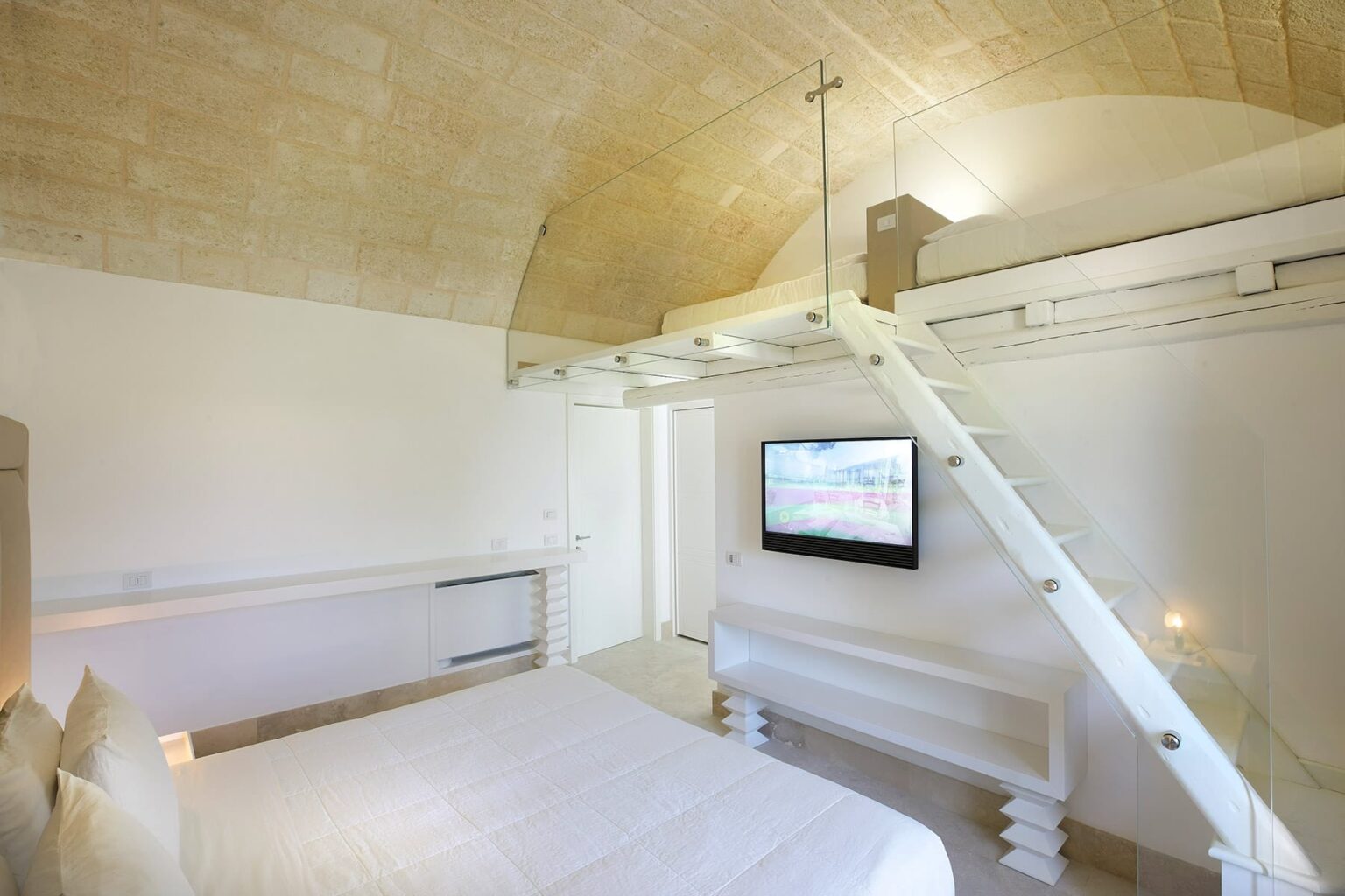 Modern minimalist bedroom with a lofted sleeping area, featuring a comfortable bed, a sleek white staircase, and a wall-mounted television. The room has a vaulted ceiling and neutral tones, creating a serene and spacious atmosphere. Ideal for relaxation and contemporary living. | Only on SopranoVillas