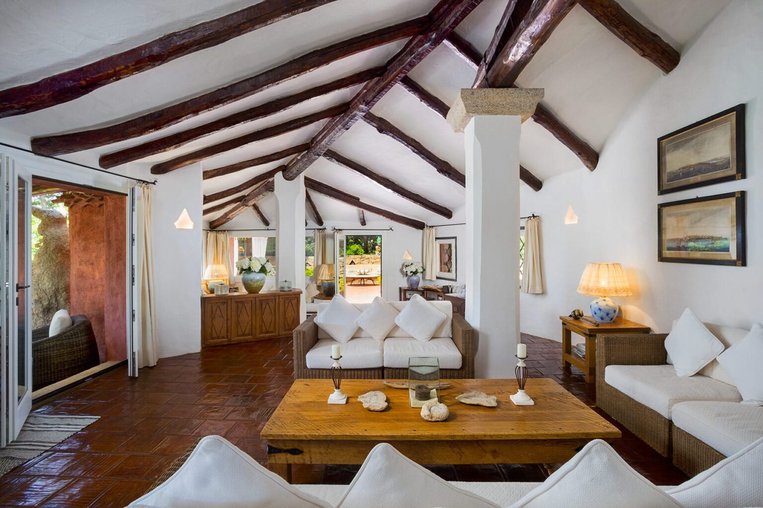 Spacious living room with high wooden beams, white walls, and cozy furniture, featuring a wooden coffee table, soft sofas, and decorative art. Natural light floods the space through large windows, creating a warm and inviting atmosphere. | Only on SopranoVillas