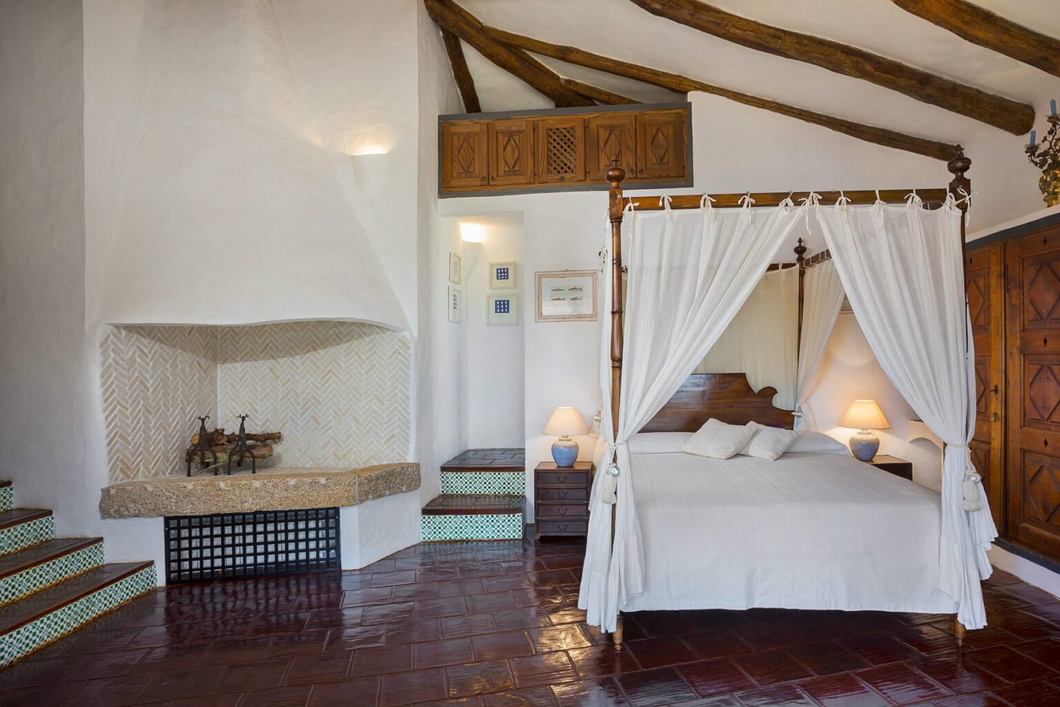 Cozy bedroom with a four-poster bed draped in white curtains, featuring a rustic fireplace and wooden beam ceiling. The room includes stairs leading to an upper area and warm lighting for a relaxing ambiance. Ideal for a romantic getaway or peaceful retreat. | Only on SopranoVillas