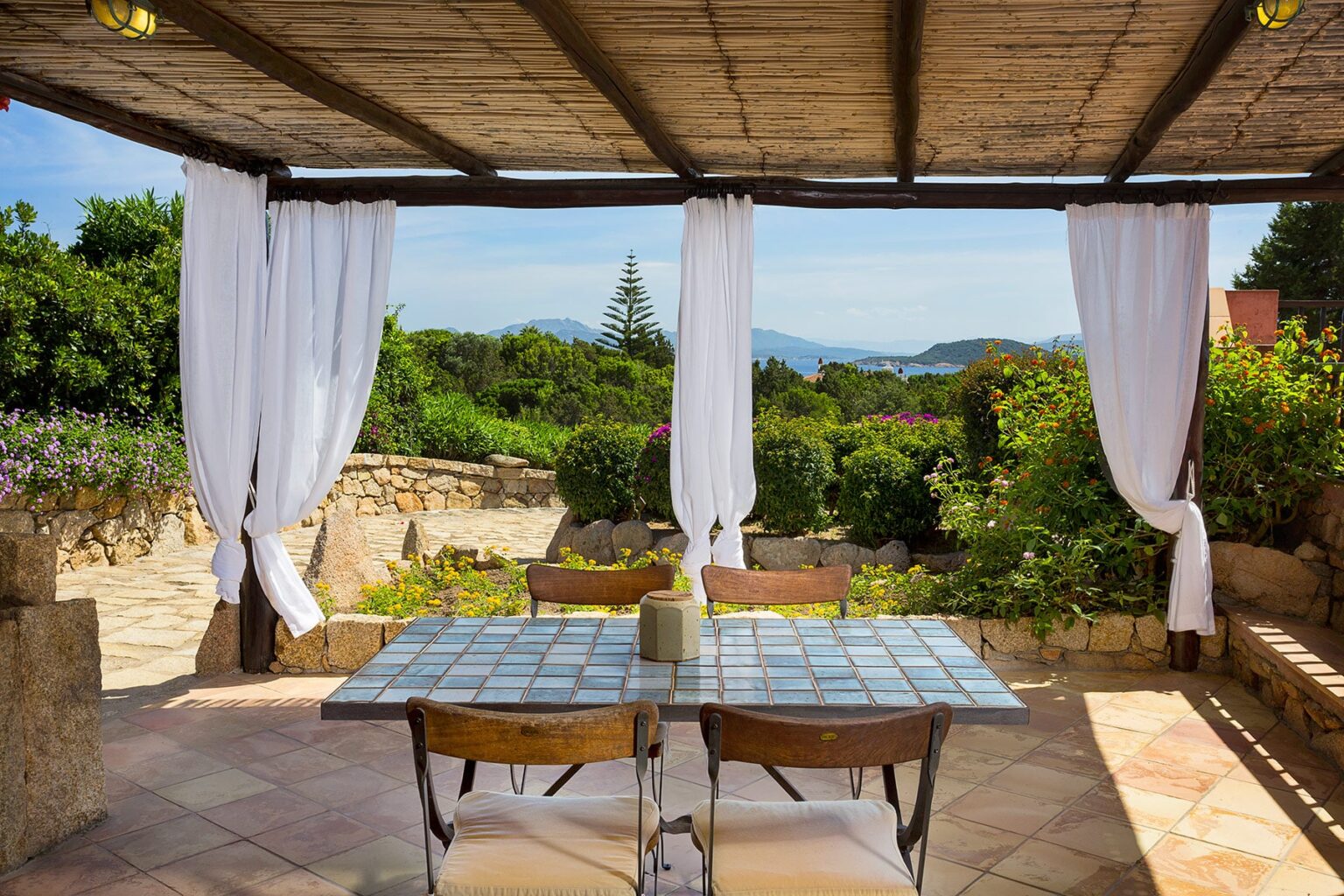 Outdoor dining area under a wooden pergola, featuring a tiled table and wooden chairs, surrounded by lush greenery and distant mountains, with white curtains gently swaying in the breeze. | Only on SopranoVillas