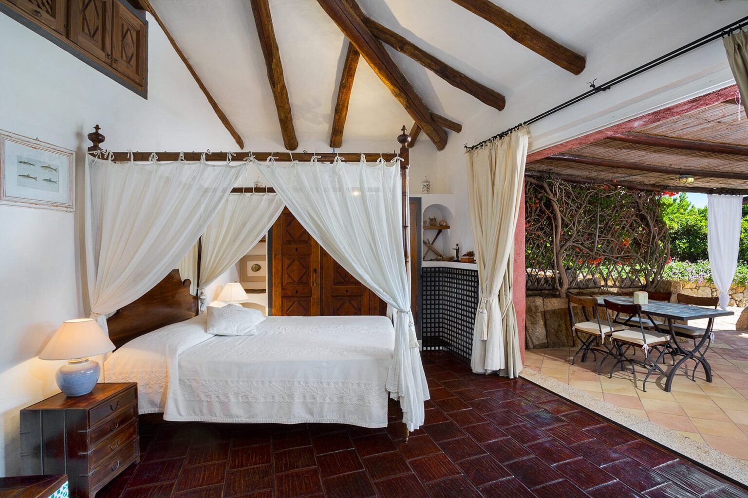 Cozy bedroom with a four-poster bed, white linens, and wooden accents, featuring exposed beam ceilings. The room opens to a patio with outdoor dining furniture, surrounded by greenery, creating a serene atmosphere. Ideal for a relaxing getaway. | Only on SopranoVillas