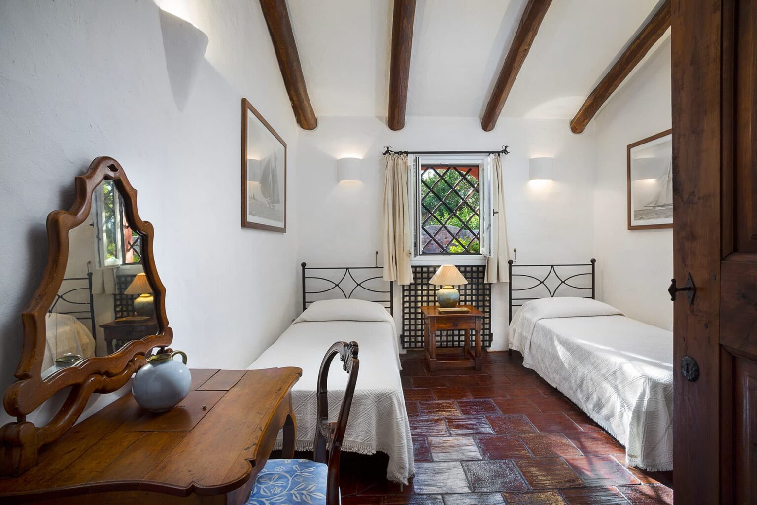 Cozy bedroom featuring two twin beds, wooden ceiling beams, a vintage mirror, and a small table with a vase. Natural light filters through a window adorned with decorative glass, enhancing the inviting atmosphere. | Only on SopranoVillas
