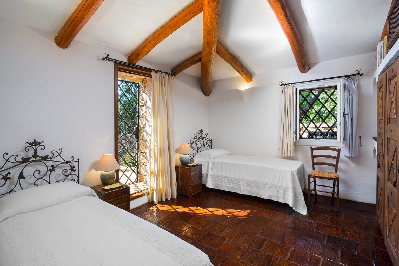 Cozy bedroom with two twin beds, wooden beams, and natural light from windows, featuring a rustic charm with iron bed frames and a wooden chair. Ideal for relaxing stays. | Only on SopranoVillas