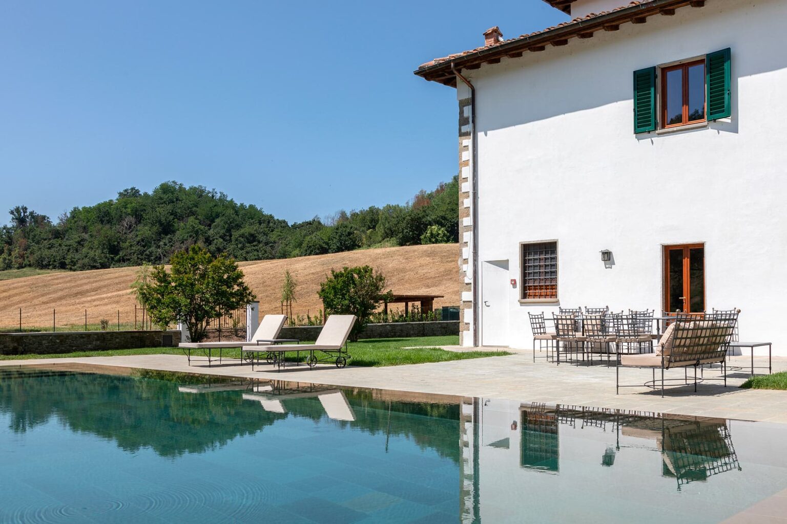 Modern white villa with green shutters, featuring a swimming pool and outdoor seating area, surrounded by lush greenery and rolling hills under a clear blue sky. Ideal for relaxation and luxury vacations. | Only on SopranoVillas