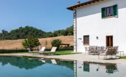 Modern white villa with green shutters, featuring a swimming pool and outdoor seating area, surrounded by lush greenery and rolling hills under a clear blue sky. Ideal for relaxation and luxury vacations.