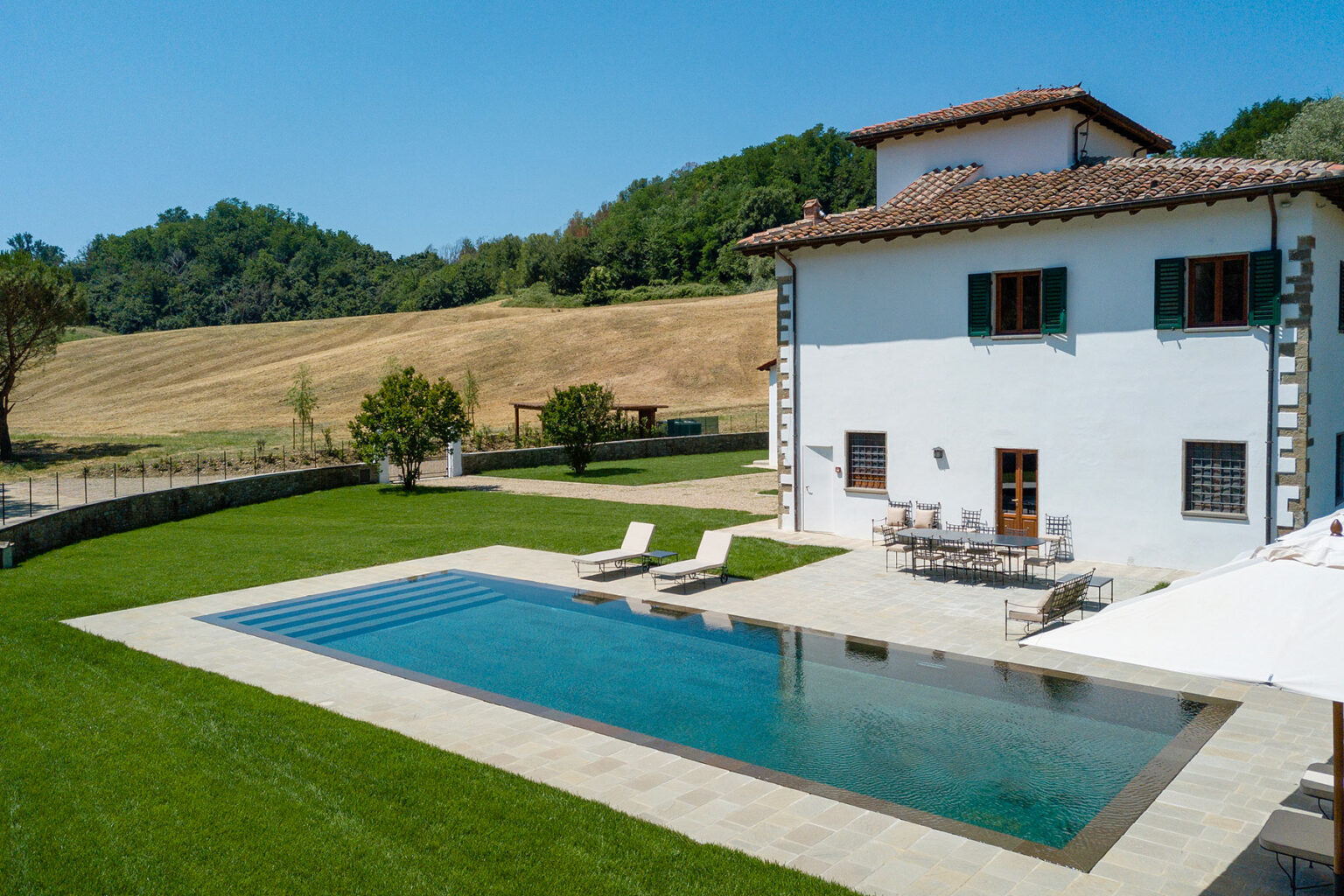 Aerial view of a modern villa with a swimming pool, surrounded by lush green lawns and gentle rolling hills in the background, ideal for vacation rentals or luxury retreats. | Only on SopranoVillas