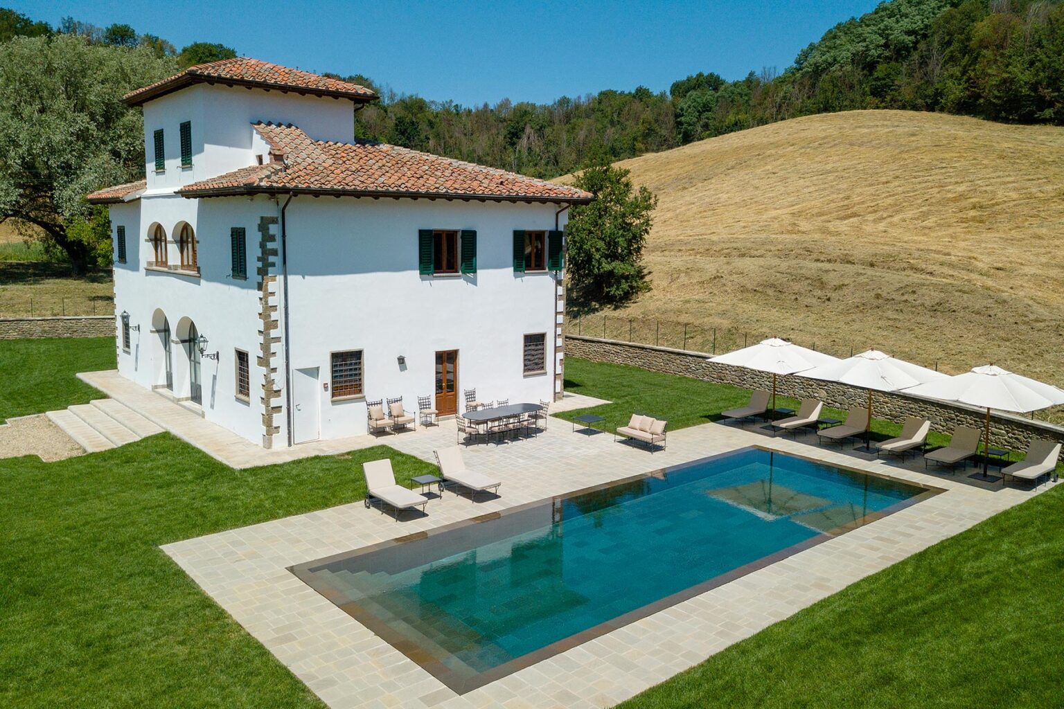 Aerial view of a charming white villa with a tile roof, surrounded by lush green grass and a serene landscape. The property features a sparkling swimming pool and several lounge chairs under shaded umbrellas, perfect for relaxation in a tranquil setting. | Only on SopranoVillas