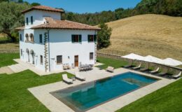Aerial view of a charming white villa with a tile roof, surrounded by lush green grass and a serene landscape. The property features a sparkling swimming pool and several lounge chairs under shaded umbrellas, perfect for relaxation in a tranquil setting.