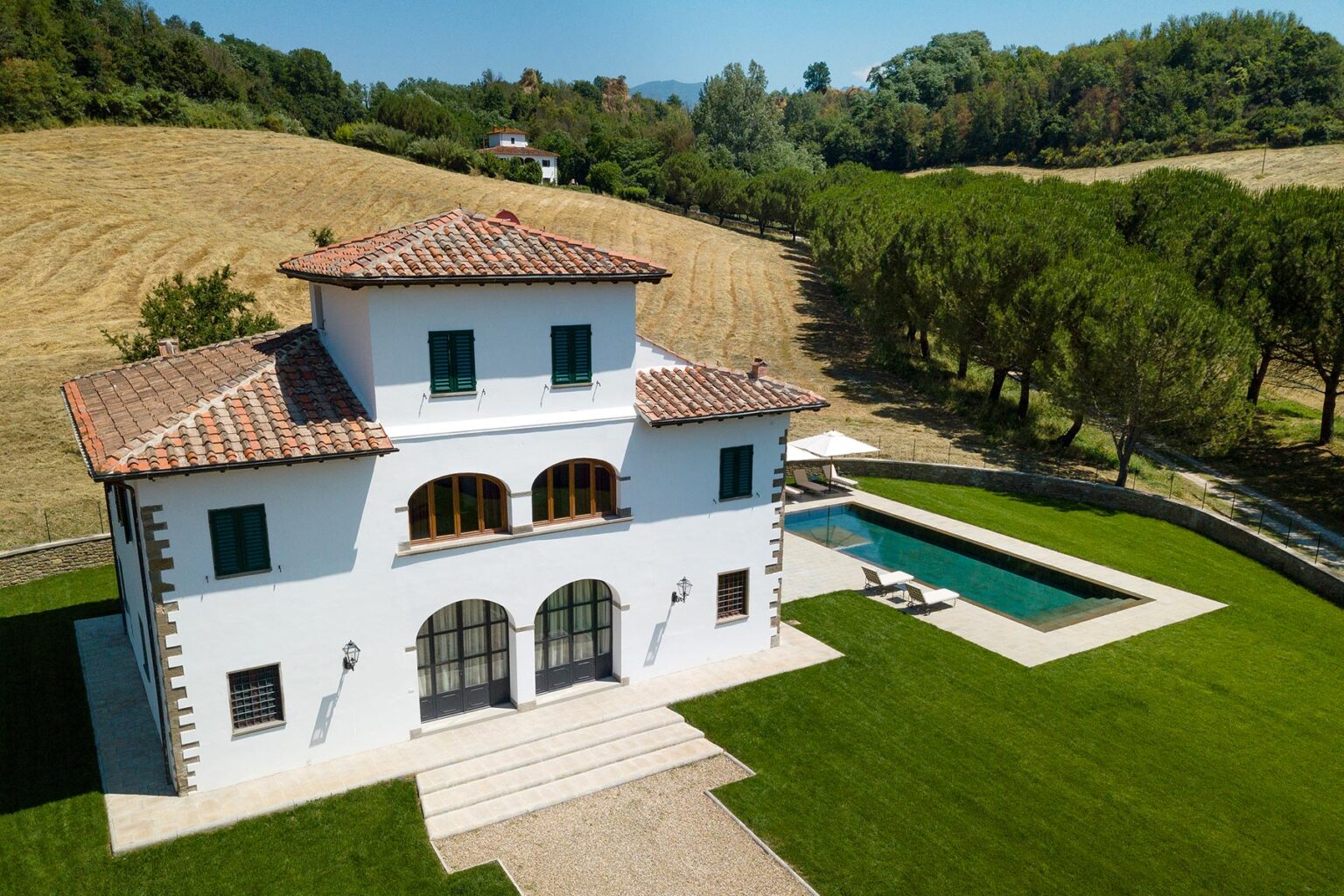 Aerial view of a modern white villa with a tiled roof, surrounded by lush green lawns and trees, featuring a swimming pool in the backyard, set against a scenic landscape. | Only on SopranoVillas