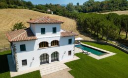 Aerial view of a modern white villa with a tiled roof, surrounded by lush green lawns and trees, featuring a swimming pool in the backyard, set against a scenic landscape.