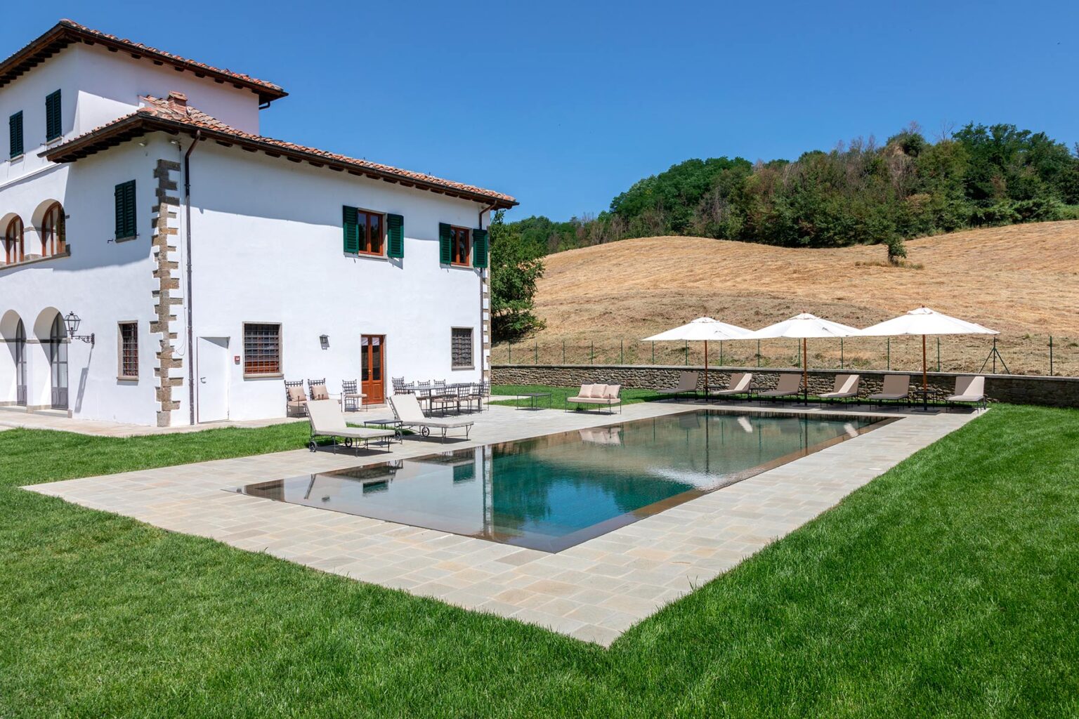 Luxury villa with a modern design featuring a swimming pool, surrounded by lush green grass and shaded lounge areas, set against a backdrop of rolling hills and blue skies. Ideal for vacation rentals or private retreats. | Only on SopranoVillas