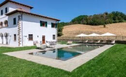 Luxury villa with a modern design featuring a swimming pool, surrounded by lush green grass and shaded lounge areas, set against a backdrop of rolling hills and blue skies. Ideal for vacation rentals or private retreats.