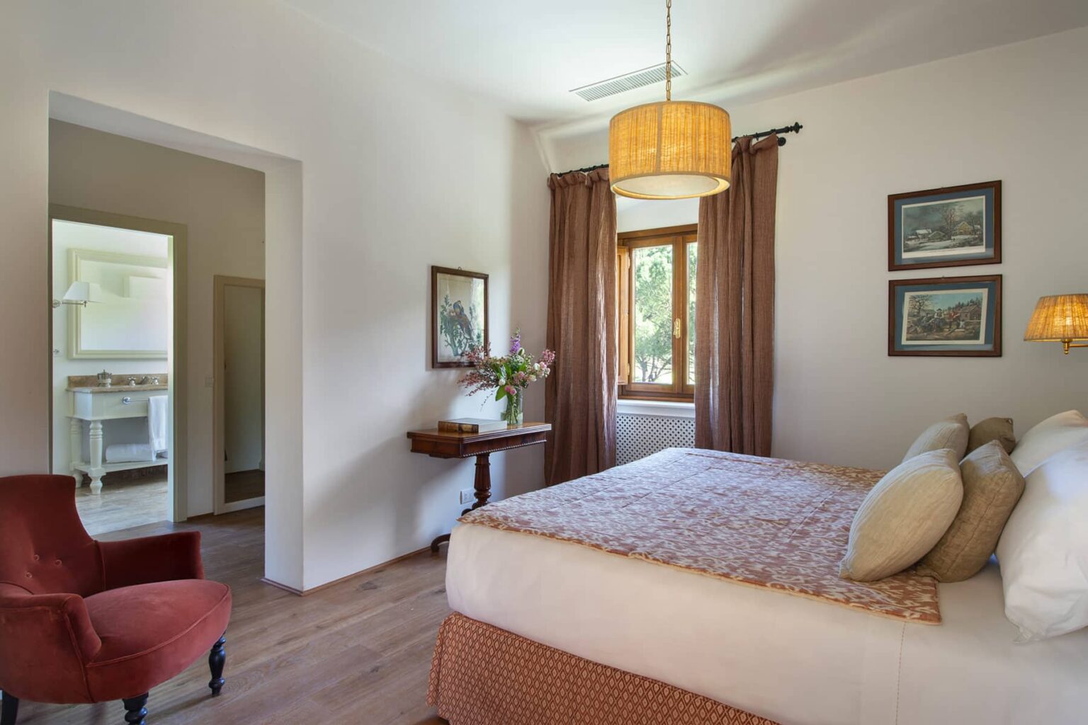 Cozy bedroom featuring a queen-sized bed with decorative pillows, elegant curtains, a stylish lamp, and a vintage armchair. Natural light floods the room through a window, enhancing the inviting atmosphere. A glimpse of a bathroom can be seen through the doorway, adding to the room's functionality. | Only on SopranoVillas