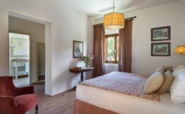 Cozy bedroom featuring a queen-sized bed with decorative pillows, elegant curtains, a stylish lamp, and a vintage armchair. Natural light floods the room through a window, enhancing the inviting atmosphere. A glimpse of a bathroom can be seen through the doorway, adding to the room's functionality.