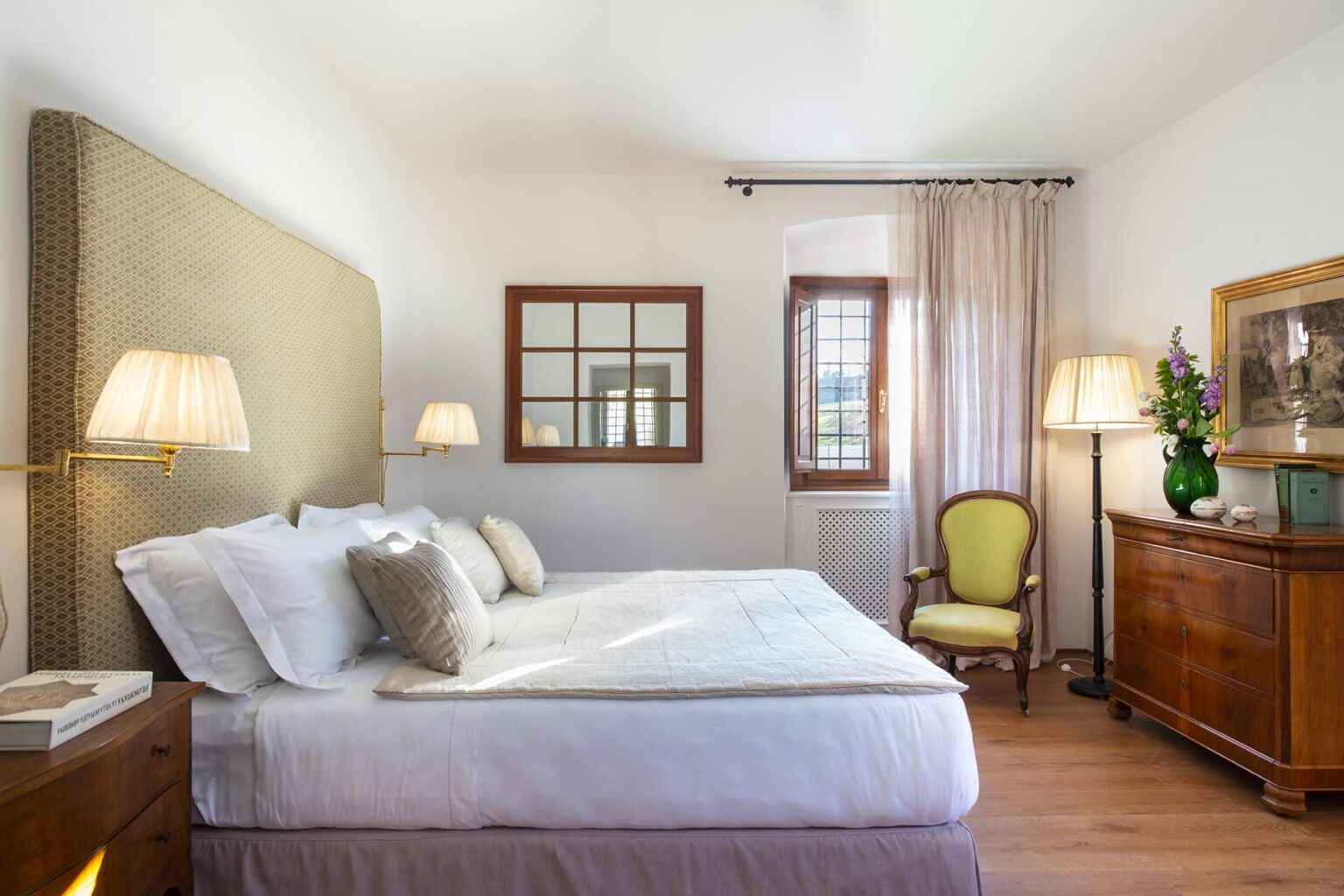 Cozy and elegantly designed bedroom featuring a comfortable bed with decorative pillows, soft lighting from bedside lamps, a vintage wooden dresser, and a green accent chair, all set against a bright, airy backdrop with natural light. | Only on SopranoVillas