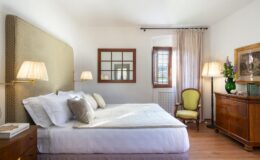 Cozy and elegantly designed bedroom featuring a comfortable bed with decorative pillows, soft lighting from bedside lamps, a vintage wooden dresser, and a green accent chair, all set against a bright, airy backdrop with natural light.