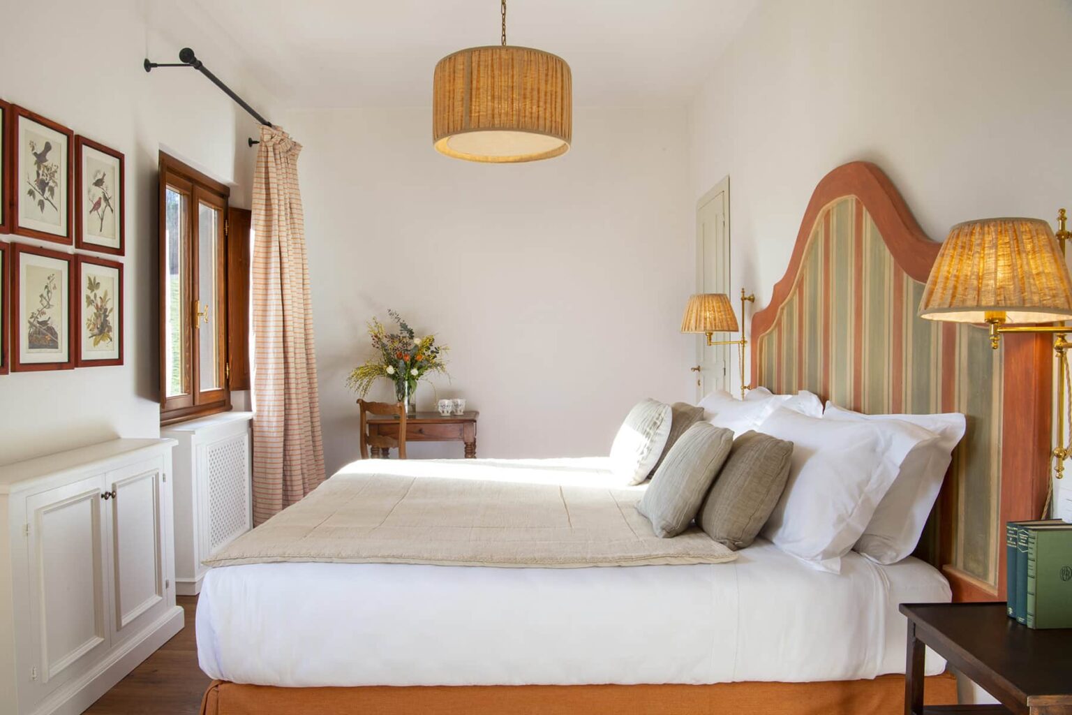 Cozy and stylish bedroom featuring a large bed with decorative pillows, a warm color palette, and elegant lighting. The room includes natural light from a window, framed artwork on the wall, and a small table with a vase of flowers, creating a serene atmosphere. | Only on SopranoVillas