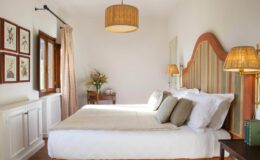 Cozy and stylish bedroom featuring a large bed with decorative pillows, a warm color palette, and elegant lighting. The room includes natural light from a window, framed artwork on the wall, and a small table with a vase of flowers, creating a serene atmosphere.