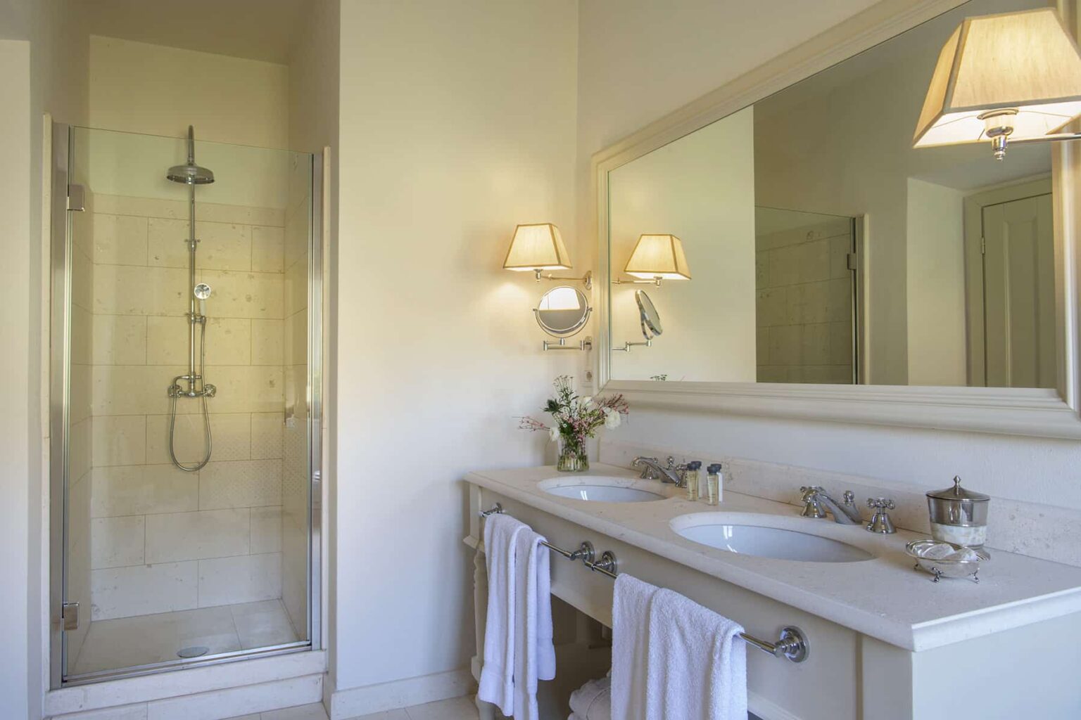 Modern bathroom featuring a glass shower enclosure, double vanity with sinks, elegant wall sconces, and a large mirror. The space is bright and airy, enhanced by neutral colors and decorative touches like fresh flowers and neatly arranged towels. | Only on SopranoVillas