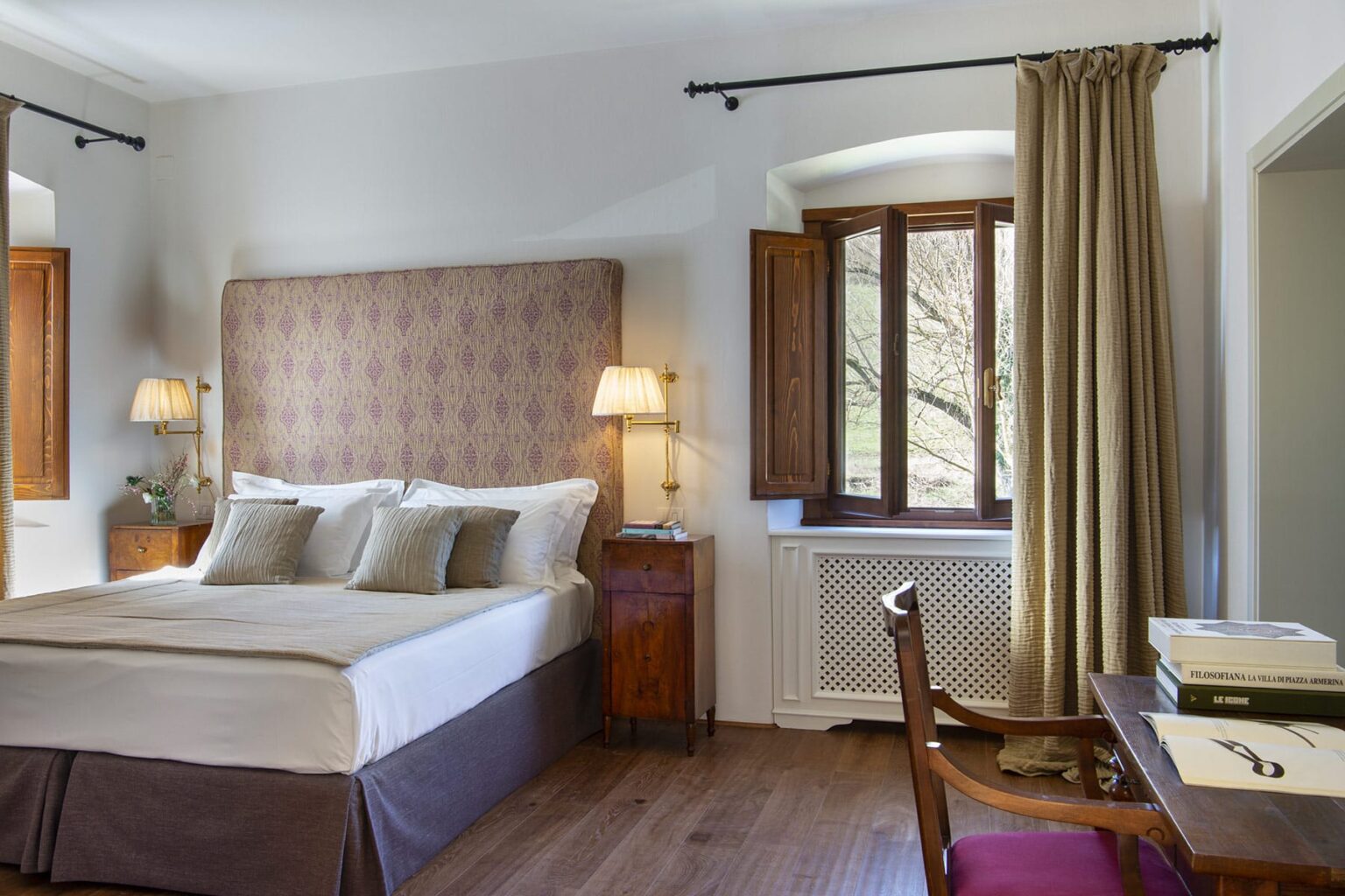 Cozy hotel room featuring a queen-sized bed with elegant bedding, wooden furniture, and large windows dressed with beige curtains, providing natural light and a warm atmosphere. | Only on SopranoVillas