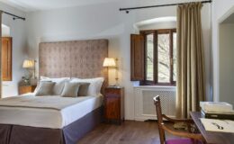 Cozy hotel room featuring a queen-sized bed with elegant bedding, wooden furniture, and large windows dressed with beige curtains, providing natural light and a warm atmosphere.