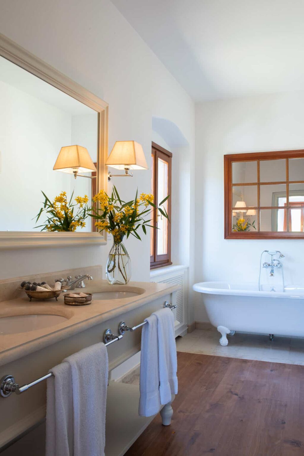 Elegant bathroom featuring a freestanding bathtub, double sinks, and a large mirror. The decor includes stylish lamps and a vase of fresh yellow flowers, enhancing the bright and airy atmosphere. Natural light streams in through the windows, creating a serene and inviting space. | Only on SopranoVillas