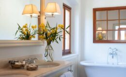 Elegant bathroom featuring a freestanding bathtub, double sinks, and a large mirror. The decor includes stylish lamps and a vase of fresh yellow flowers, enhancing the bright and airy atmosphere. Natural light streams in through the windows, creating a serene and inviting space.