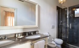 Modern bathroom featuring dual sinks, a large mirror, and a glass shower enclosure with contemporary fixtures, complemented by warm lighting and neutral tones.