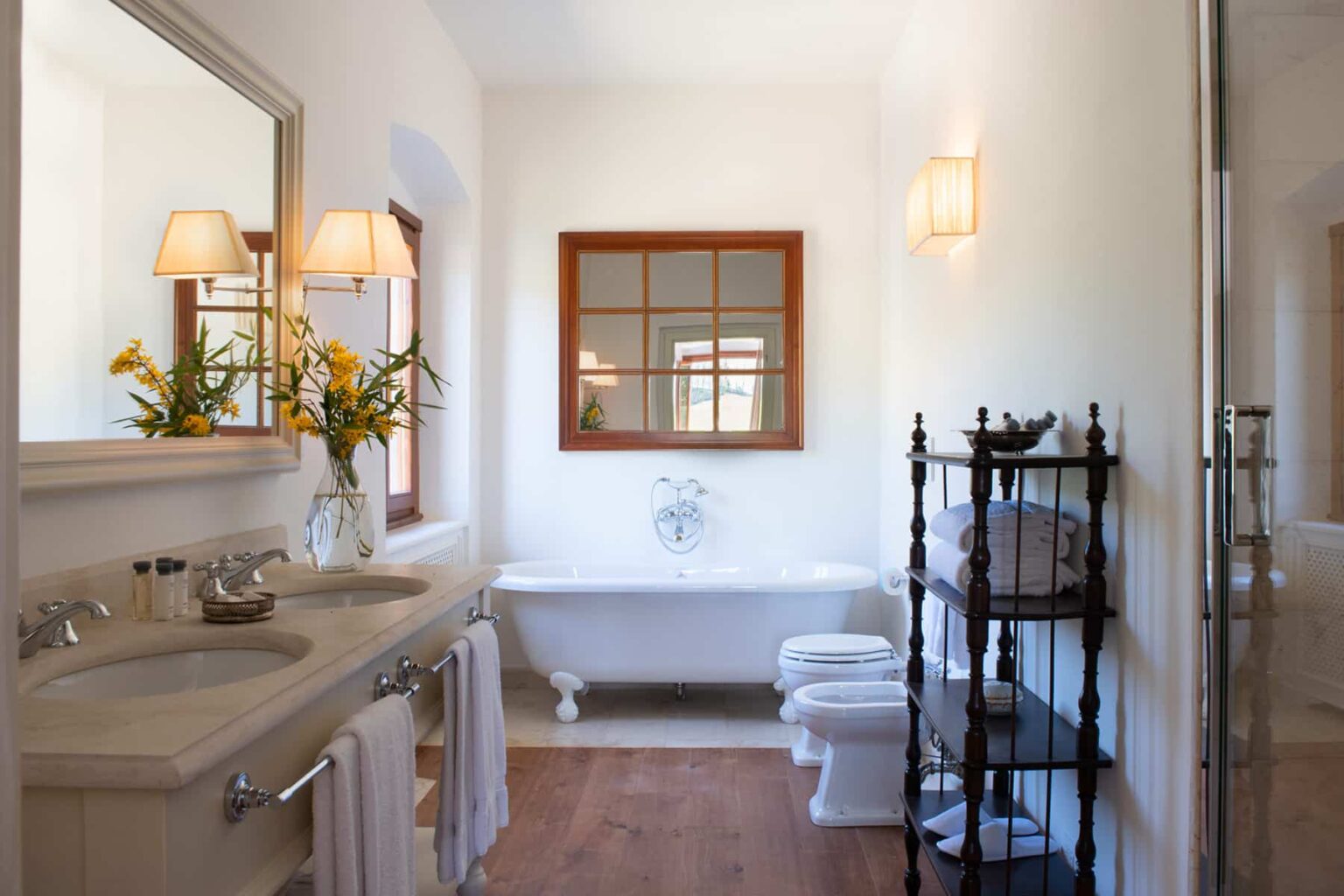 Bright and spacious bathroom featuring a double vanity, large mirror, and elegant fixtures. A freestanding bathtub is paired with a stylish window and decorative lighting, complemented by a towel rack and fresh flowers for a touch of elegance. | Only on SopranoVillas