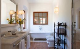 Bright and spacious bathroom featuring a double vanity, large mirror, and elegant fixtures. A freestanding bathtub is paired with a stylish window and decorative lighting, complemented by a towel rack and fresh flowers for a touch of elegance.