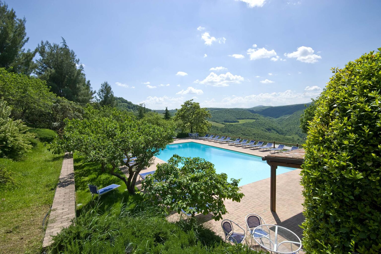 Scenic view of a tranquil swimming pool surrounded by lush greenery and hills under a clear blue sky, ideal for relaxation and outdoor leisure activities. | Only on SopranoVillas