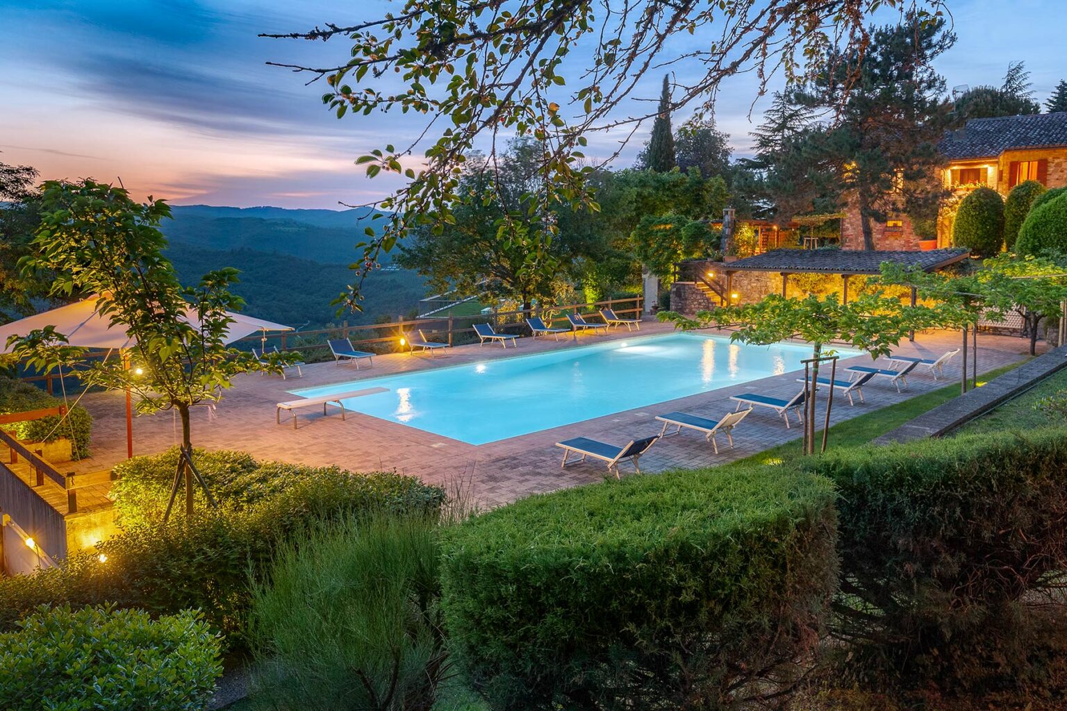 Scenic view of a luxurious outdoor swimming pool surrounded by lush greenery and illuminated by soft lighting, set against a backdrop of rolling hills and a colorful sunset sky. | Only on SopranoVillas