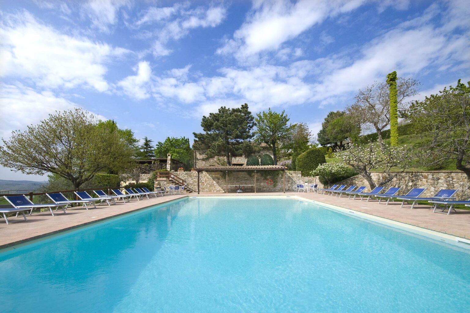 Outdoor swimming pool surrounded by lounge chairs and lush greenery, with a clear blue sky and scattered clouds in the background. Ideal for relaxation and leisure activities. | Only on SopranoVillas