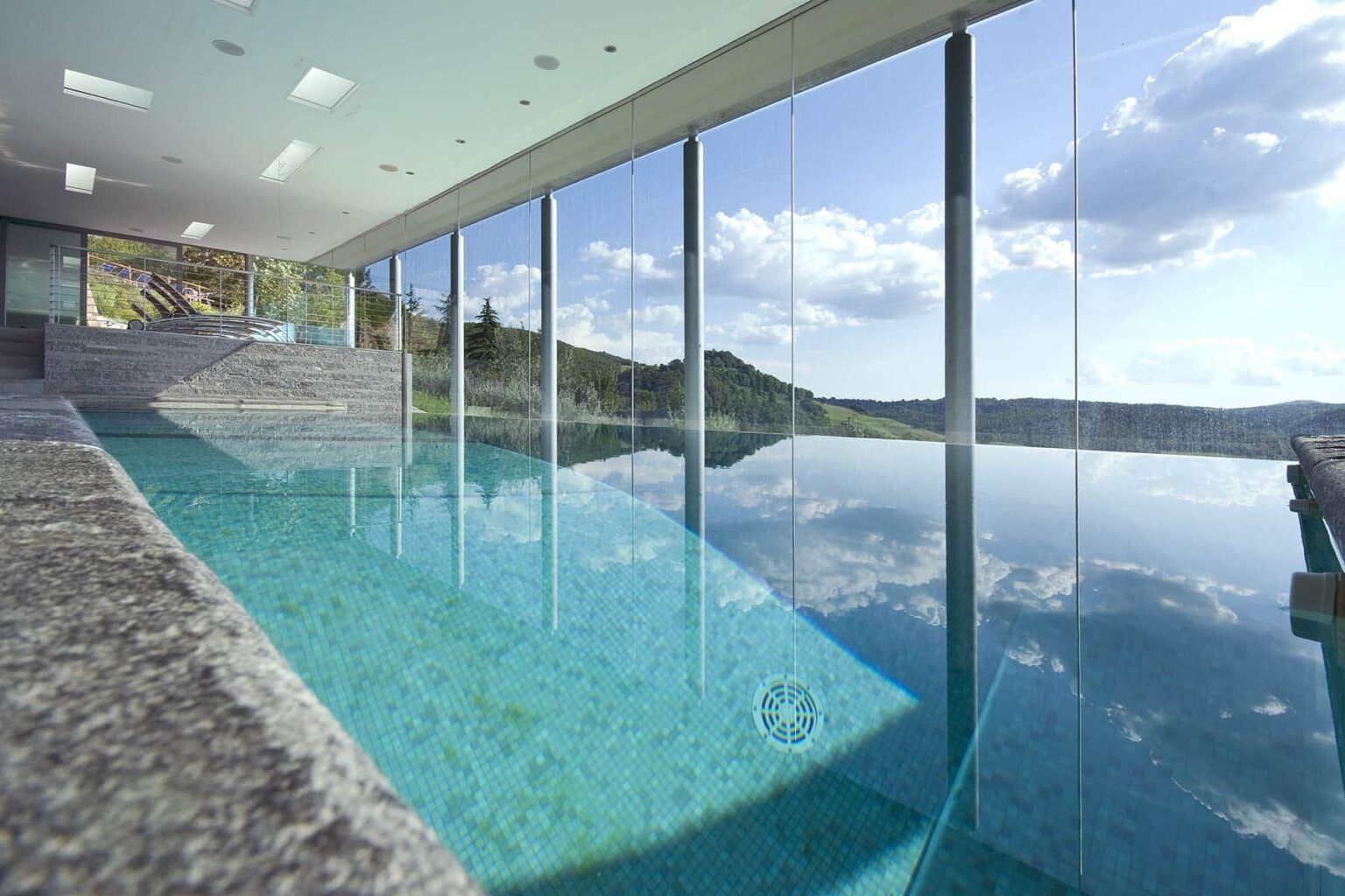 Modern indoor swimming pool with floor-to-ceiling glass walls, offering panoramic views of a serene landscape and a clear blue sky. The pool features a sleek design with a stone edge, reflecting the surrounding nature. | Only on SopranoVillas