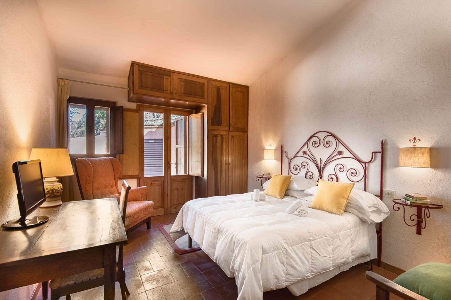 Cozy hotel room featuring a double bed with yellow accent pillows, a wooden desk, and large windows allowing natural light. The room has warm wooden accents and a comfortable seating area, ideal for relaxation. | Only on SopranoVillas